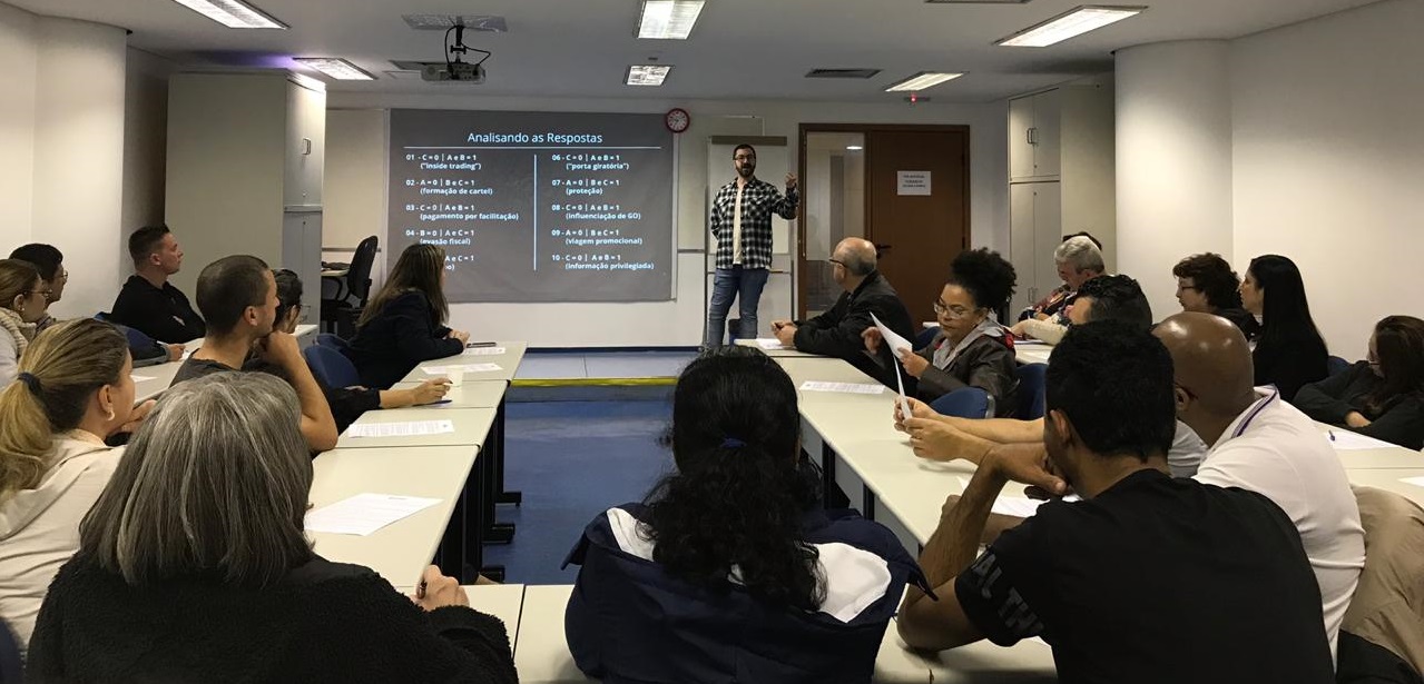 Foto do instrutor ao fundo, e dos alunos sentados nas cadeiras e prestando atenção na aula.